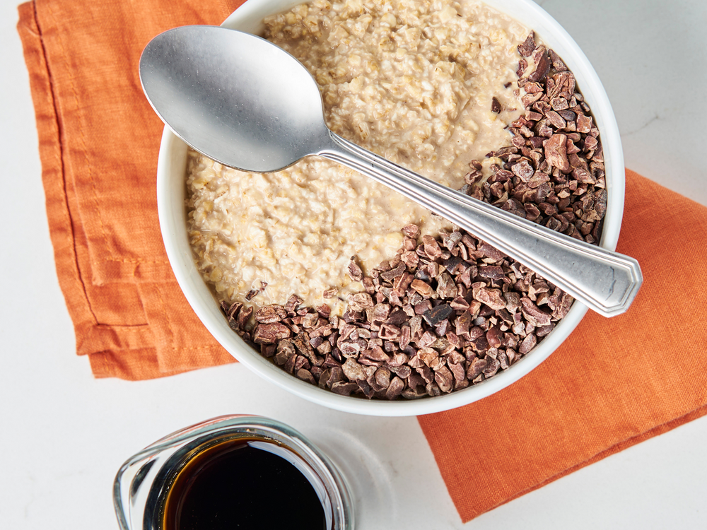 White bowl with oatmeal and cacao nibs on an orange napkin, with a spoon and small jar of syrup.
